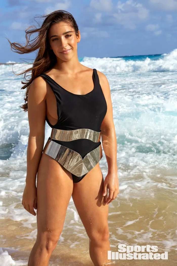 Aly Raisman smiles in a black one-piece and silver metallic belt in front of the crashing waves.